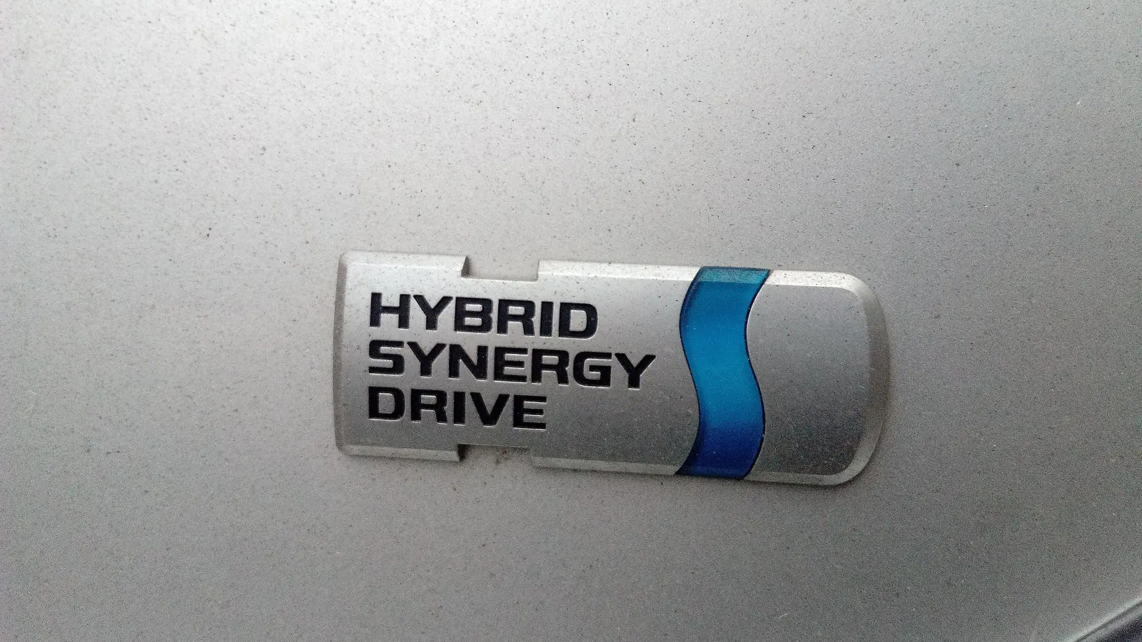Gros plan sur l’emblème chromé Hybrid Synergy Drive de Toyota, sur une carrosserie grise